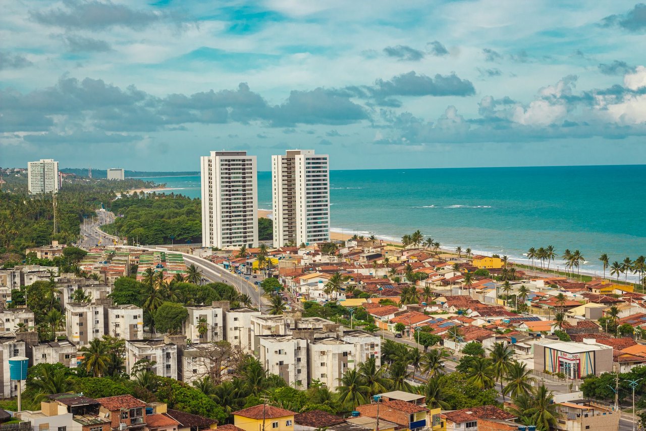 Vista urbana de Maceió com edifícios residenciais, áreas verdes e a faixa litorânea ao fundo.