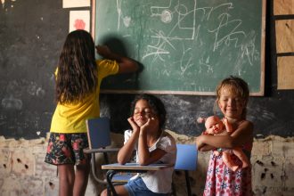Três crianças brincam e aprendem diante de um quadro-negro em uma sala de aula.